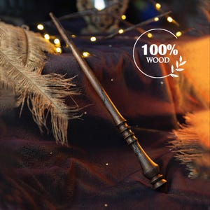 Op de afbeelding: Een donkerbruine houten toverstaf met ingewikkelde details en een zwart en bruin swirl-patroon. De toverstaf ligt op een donkerpaarse stof met gouden accenten. De tekst "100% HOUT" is zichtbaar in een witte cirkel.