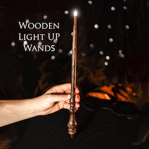 Op de afbeelding: Een houten toverstaf met een gloeiende punt wordt tegen een donkere achtergrond gehouden met de tekst "Wooden Light Up Wands". De toverstaf heeft een gedetailleerde, gesneden handgreep en een slanke, taps toelopende body. De punt straalt een zacht, wit licht uit, wat een magisch effect creëert.