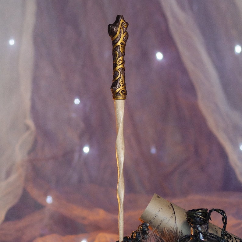May include: A wooden wand with a twisted, light brown shaft and a gold-painted, detailed, brown handle. The wand is standing upright against a blurry background of white fabric.