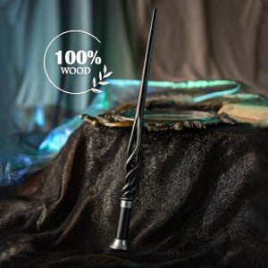 Op de afbeelding: Een zwarte houten toverstaf met een gedraaid handvat en een gladde, taps toelopende schacht. De basis van het handvat is voorzien van een zilverkleurige metalen ring en een uitlopende zilverkleurige knop. De tekst 100% WOOD staat in het wit.