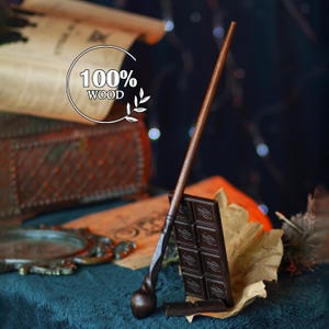 Op de afbeelding: Een donkerbruine houten toverstaf met een getextureerde handgreep en een afgerond uiteinde, leunend tegen een reep pure chocolade. De afbeelding bevat de tekst "100% WOOD", wat het materiaal van de toverstaf aangeeft.