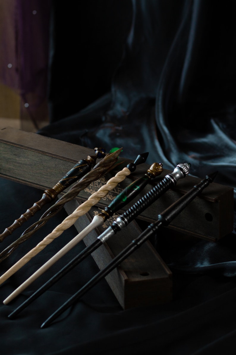May include: A set of six wooden wands with different colored tips and designs. The wands are arranged in a wooden box with a black velvet background.