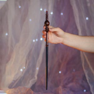 May include: A wooden wand with a dark brown handle and a black shaft. The handle has intricate carved details and a red tip. The wand is held in a person's hand against a soft, blurred background with bokeh lights.