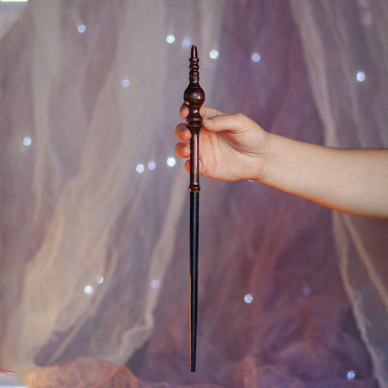 May include: A wooden wand with a dark brown handle and a black shaft. The handle has intricate carved details and a red tip. The wand is held in a person's hand against a soft, blurred background with bokeh lights.
