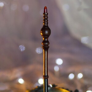 May include: A dark brown, ornate wand with a red, rounded tip. The wand has a detailed, carved design with a bulbous center and a slender handle. The background is blurred with soft, out-of-focus lights.
