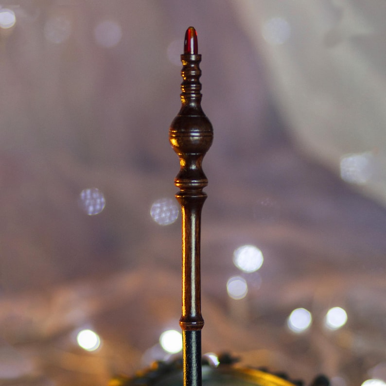 May include: A dark brown, ornate wand with a red, rounded tip. The wand has a detailed, carved design with a bulbous center and a slender handle. The background is blurred with soft, out-of-focus lights.