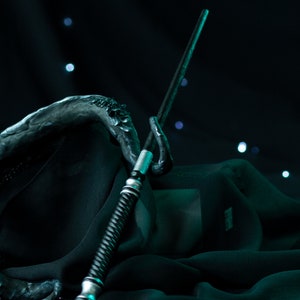 May include: A silver and black wand with a twisted handle rests on a dark green fabric. The wand is pointed upwards and the fabric is draped around it.