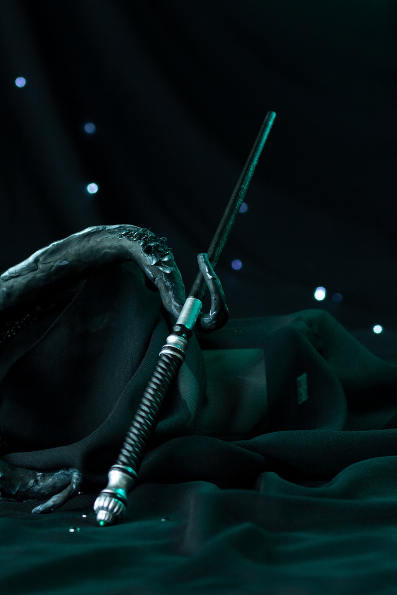 May include: A silver and black wand with a twisted handle rests on a dark green fabric. The wand is pointed upwards and the fabric is draped around it.