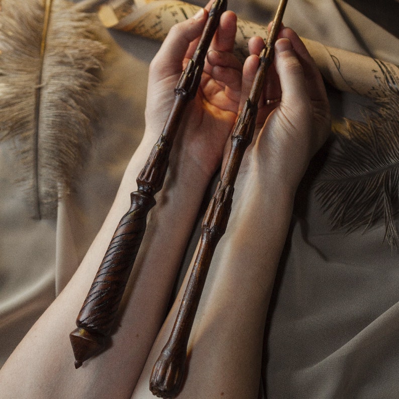 May include: Two wooden wands with intricate details, held by hands against a brown background.