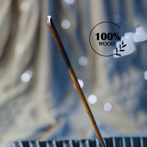 Puede incluir: Una varita de madera con punta transparente, con un mango marrón oscuro que hace la transición a un marrón más claro. La varita se encuentra sobre un fondo borroso con el texto "100% WOOD" en un círculo negro.