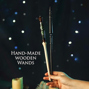 Op de afbeelding: Twee handgemaakte houten toverstokken. Eén is zwart met een punt. De andere heeft een licht houten handvat, een groen en bruin patroon in het midden en een decoratieve bovenkant. De tekst "HAND-MADE WOODEN WANDS" wordt weergegeven.