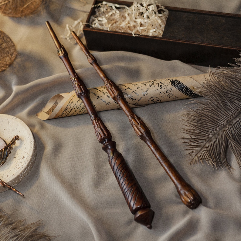 May include: Two wooden wands with intricate details, lying on a brown fabric surface with a rolled up piece of paper and a feather. The paper has writing on it, including the word "Sectilum".