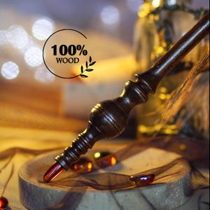 May include: A dark brown wooden wand with intricate carvings and a red crystal tip. The wand is labeled "100% WOOD" and is set against a blurred background with warm lighting. The wand is a magical item.