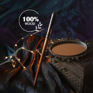 Op de afbeelding: Een houten toverstaf met een donkere metalen punt en accenten. De toverstaf is bruin met een donkere metalen punt en accenten. De afbeelding bevat een decoratieve spiegel en de tekst "100% WOOD". De achtergrond is donker met lichtjes.