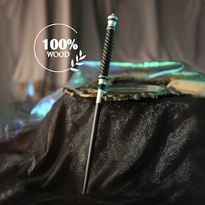 May include: A black wooden wand with a silver handle and a twisted black grip. The wand has a small green gem at the top. The text "100% WOOD" is in a white circle. The wand is a prop or collectible.