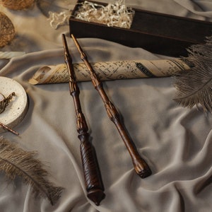 May include: Three wooden wands with intricate details lie on a brown fabric surface. The wands are brown with a dark brown finish. The fabric has a soft, wrinkled texture.