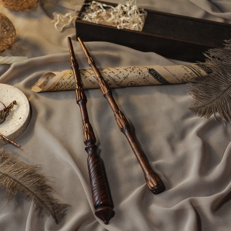 May include: Three wooden wands with intricate details lie on a brown fabric surface. The wands are brown with a dark brown finish. The fabric has a soft, wrinkled texture.