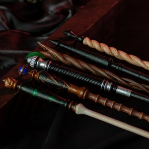 May include: A collection of six wooden wands with various designs and colors, including black, brown, green, and white. The wands are arranged on a dark surface.
