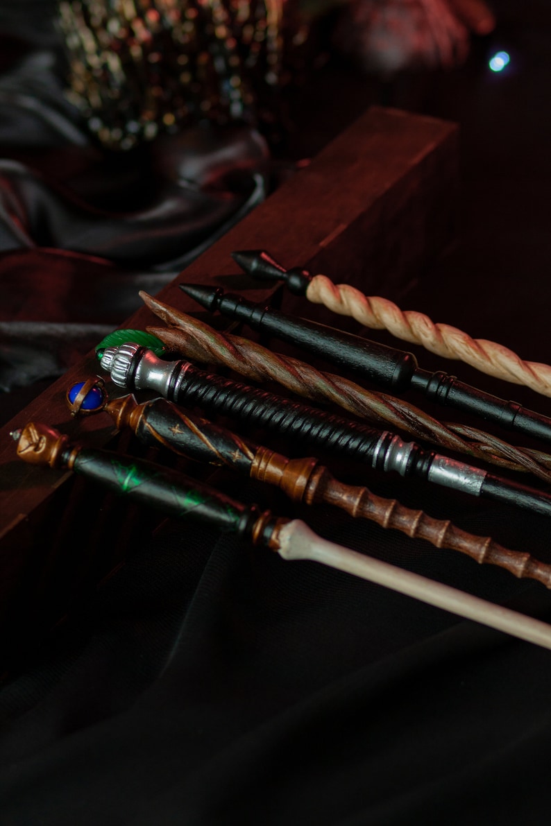 May include: A collection of six wooden wands with various designs and colors, including black, brown, green, and white. The wands are arranged on a dark surface.