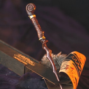 Peut inclure: Une baguette en bois brun foncé avec une spirale dorée en haut et une pointe pointue. La baguette repose sur un morceau de parchemin roulé avec le mot "HOOK" visible.
