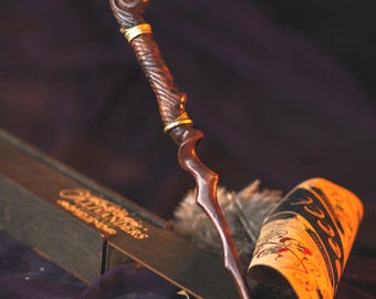 May include: A dark brown wooden wand with a gold-colored spiral at the top and a pointed tip. The wand is resting on a rolled-up piece of parchment paper with the word "HOOK" visible.