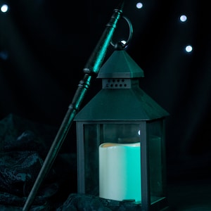May include: A black metal lantern with a glass panel and a white candle inside. The lantern is sitting on a black lace fabric with a wooden stick leaning against it. The background is a dark blue with white lights.