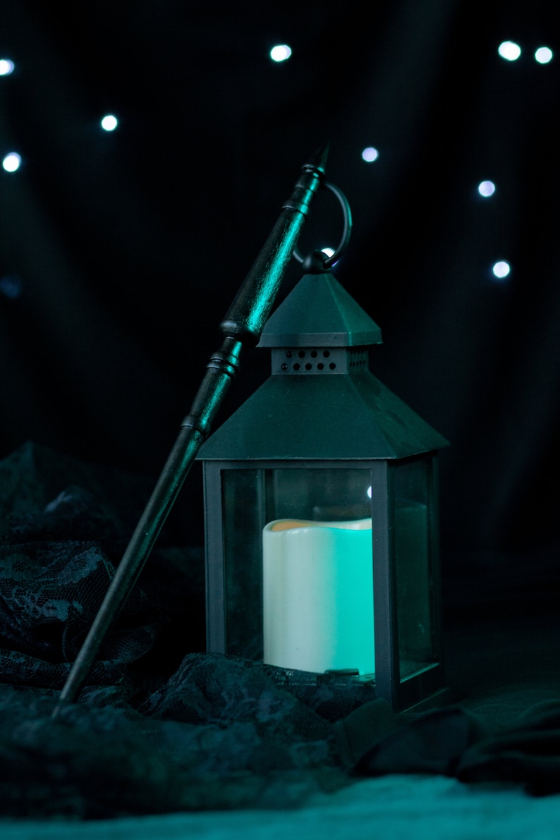 May include: A black metal lantern with a glass panel and a white candle inside. The lantern is sitting on a black lace fabric with a wooden stick leaning against it. The background is a dark blue with white lights.