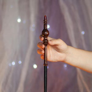 May include: A dark brown wooden wand with a bulbous handle and a slender shaft. The top of the wand features a reddish-brown crystal. The wand is held in a hand against a soft, blurred background with bokeh lights.