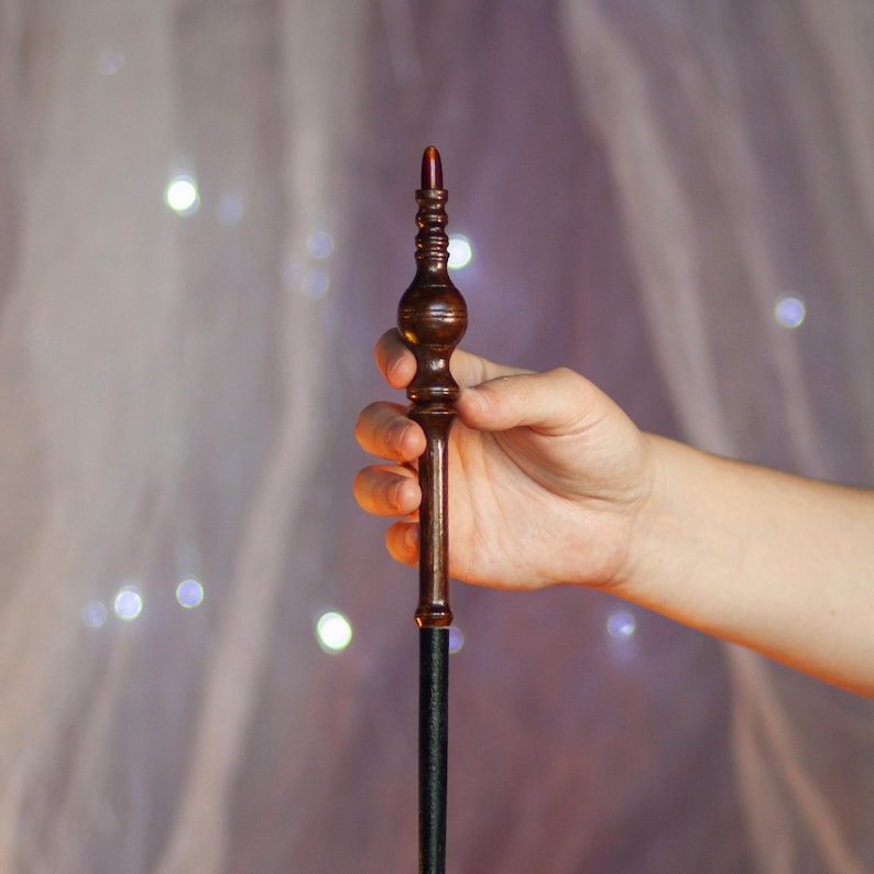 May include: A dark brown wooden wand with a bulbous handle and a slender shaft. The top of the wand features a reddish-brown crystal. The wand is held in a hand against a soft, blurred background with bokeh lights.