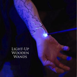 Puede incluir: Una varita mágica de madera azul con luz se apunta a la muñeca de una persona, iluminando un diseño blanco brillante que se asemeja a un rayo. El texto "LIGHT-UP WOODEN WANDS" se muestra en letras blancas.
