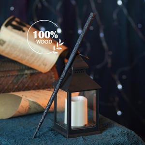 Puede incluir: Una varita de madera oscura con intrincadas tallas, apoyada contra una linterna negra con una vela blanca en su interior. La imagen incluye el texto "100% WOOD" en un círculo blanco, lo que sugiere el material de la varita. El fondo es azul oscuro con luces suaves y desenfocadas.