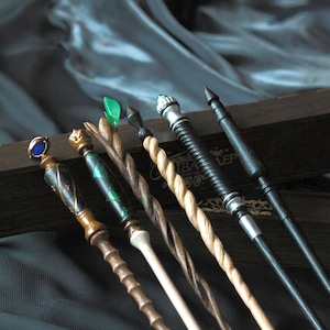May include: Six wooden wands with different coloured tips and designs. The wands are arranged in a row on a dark surface. The wands are all about 30 cm long.