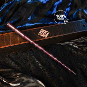 Puede incluir: Una varita de madera de color marrón rojizo oscuro con una textura segmentada similar al bambú, apoyada sobre una caja rectangular de madera oscura. La caja presenta una incrustación geométrica cuadrada y un gráfico circular con el texto 100% WOOD.