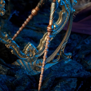 May include: A pair of wooden wands with blue and gold accents, resting on a gold ornate mirror and a black lace fabric. The wands have a dark wood finish with gold and blue details.