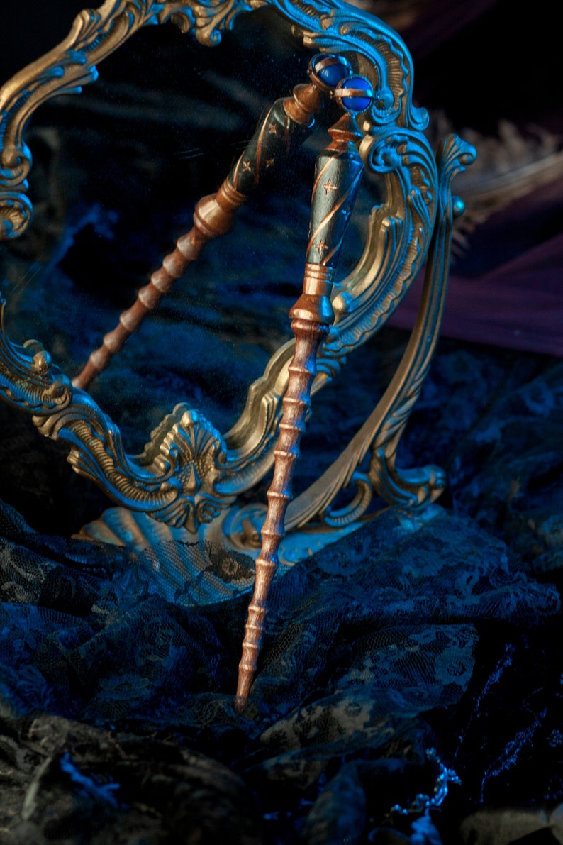 May include: A pair of wooden wands with blue and gold accents, resting on a gold ornate mirror and a black lace fabric. The wands have a dark wood finish with gold and blue details.