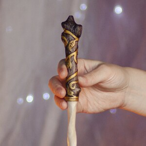 May include: A wooden wand with a brown and gold design. The wand is decorated with a vine and leaf pattern. The wand is held in a hand.