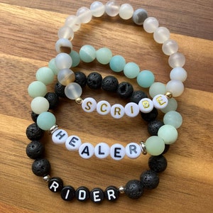 Pode incluir: Quatro pulseiras de contas com diferentes combinações de pedras e contas. Uma pulseira diz "SCRIBE" em letras douradas, outra diz "HEALER" e a última diz "RIDER". As pulseiras apresentam contas brancas, verdes, pretas e transparentes.