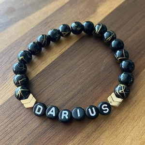 May include: A black beaded bracelet with gold detailing and the name "DARIUS" spelled out in white letters on black beads. The bracelet has gold-coloured chevron-shaped spacers and is shown on a wooden surface.