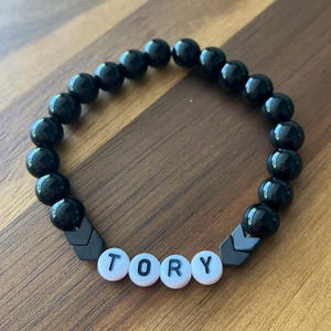May include: Black beaded bracelet with the name "TORY" spelled out in white letter beads. The bracelet features black beads and dark grey chevron-shaped accents.