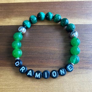 Op de afbeelding: Een armband met groene en zilveren kralen, met de naam "DRAMIONE" gespeld met zwarte letterkralen. De armband is gemaakt van ronde, gepolijste kralen. De armband wordt gepresenteerd op een houten oppervlak.
