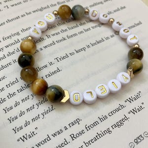 May include: A beaded bracelet with white and gold letter beads spelling "YOU" and "LOYALTY". The bracelet also features tiger eye and gold-colored beads. The bracelet is resting on an open book.