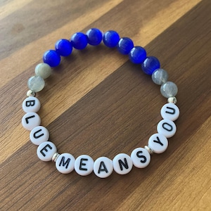 May include: A bracelet featuring blue and grey beads, with white letter beads spelling "BLUE MEANS YOU". The bracelet is displayed on a wooden surface.