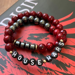 May include: Two beaded bracelets. One bracelet features dark red beads and metallic grey beads. The other bracelet has dark red beads and black beads with the words "HOUSE MARS" spelled out in white letters.
