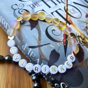 May include: A beaded bracelet featuring translucent, iridescent yellow and purple beads interspersed with white spherical beads. The white beads display the gold-lettered text DAWNDRINKER. The bracelet includes gold-toned metal accents and a small charm.