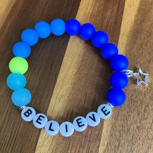 May include: A stretch bracelet featuring matte beads in shades of blue and a single lime green bead. The bracelet includes white letter beads spelling the word BELIEVE in black font and a silver-toned metal star charm attached to the side.