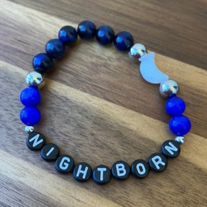 May include: A beaded bracelet featuring dark blue, bright blue, and silver-toned metallic beads. It includes a white crescent moon charm and black letter beads that spell out the word NIGHTBORN in white capital letters, arranged in a circular pattern.