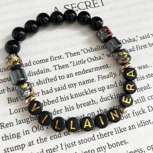 May include: Black beaded bracelet with gold lettering spelling "VILLAINE ERA". The bracelet features black, gold, and red decorative beads. The bracelet is on a book page.