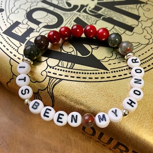 May include: A beaded bracelet with white letter beads spelling "ITS BEEN MY HONOR". The bracelet features red, brown, and green gemstone beads. The bracelet is resting on a gold-colored surface with black text.