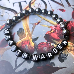 May include: A circular beaded bracelet featuring polished dark metallic hematite-style beads and black spherical beads with white printed letters spelling out the words WRAITH WARDEN. The bracelet includes small black spacers between the lettered beads.