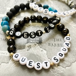 May include: Three beaded bracelets with the word "QUEST" spelled out in white letter beads. The bracelets feature black, gold, and blue beads. One bracelet spells out "QUEST SQUAD". The background is a map.